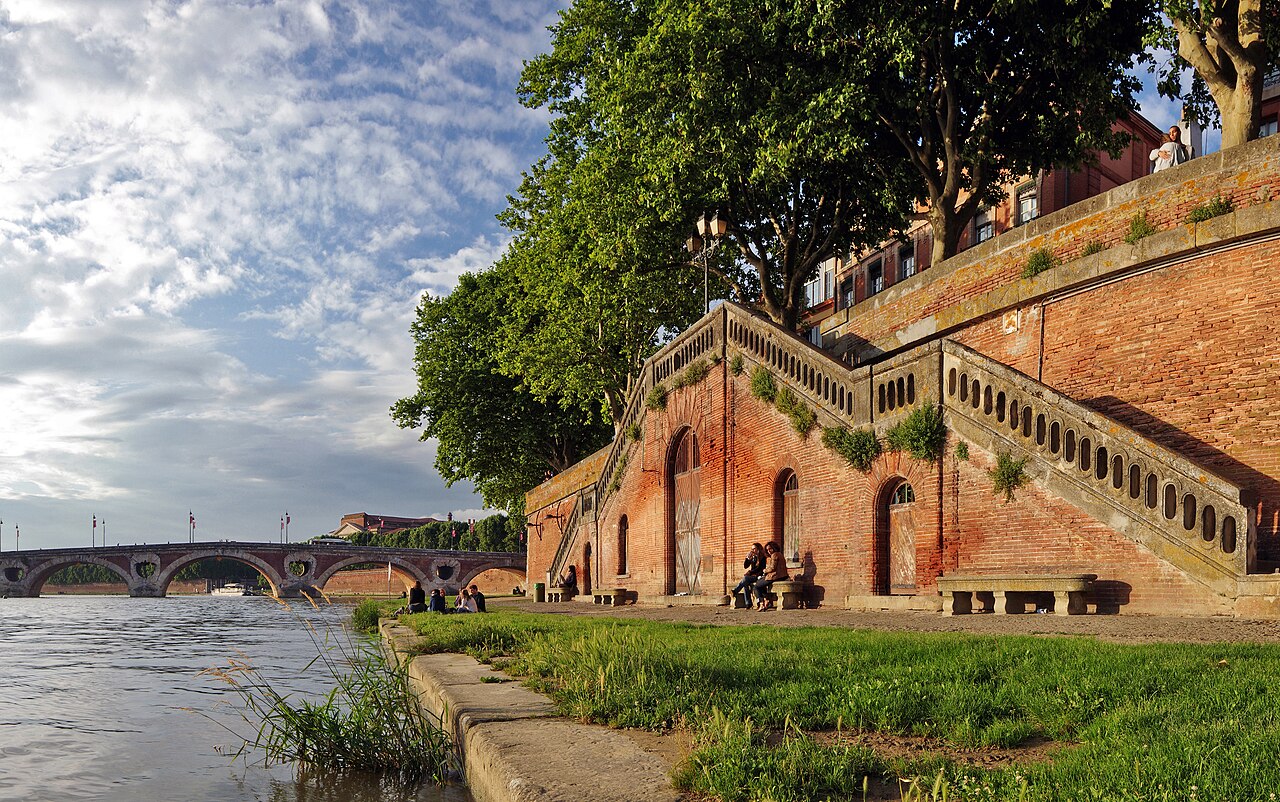 Garonne water beside a grassy bank, red-brick wall, steps and trees, with Pont Neuf in the distance.