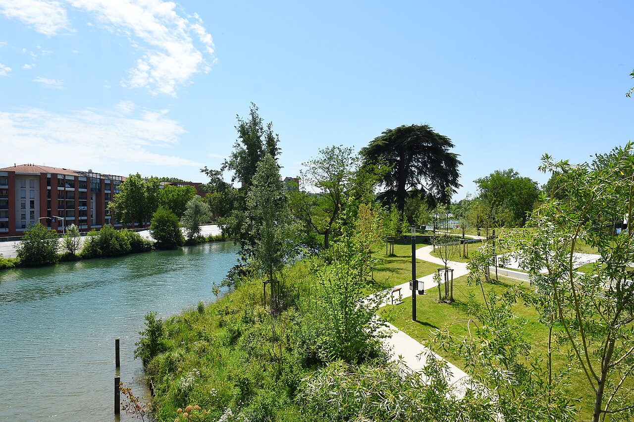 Garonne water bordered by dense green vegetation, a public path, lamps and urban buildings.