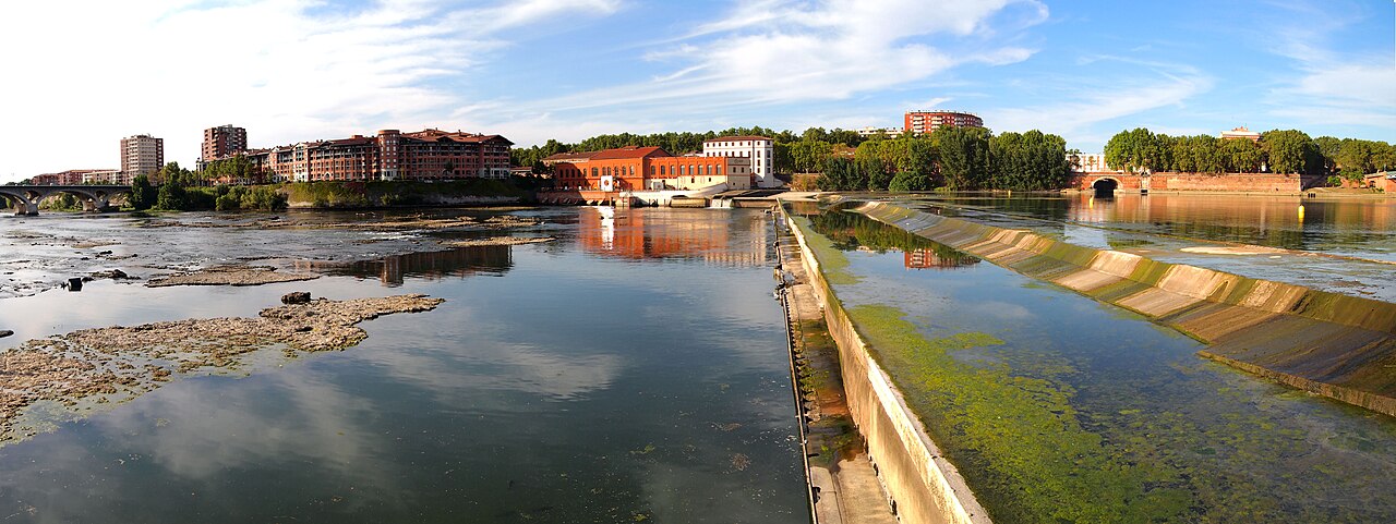 Panoramic river scene with a weir, exposed textured surfaces, reflections, bridges and buildings on the opposite bank.