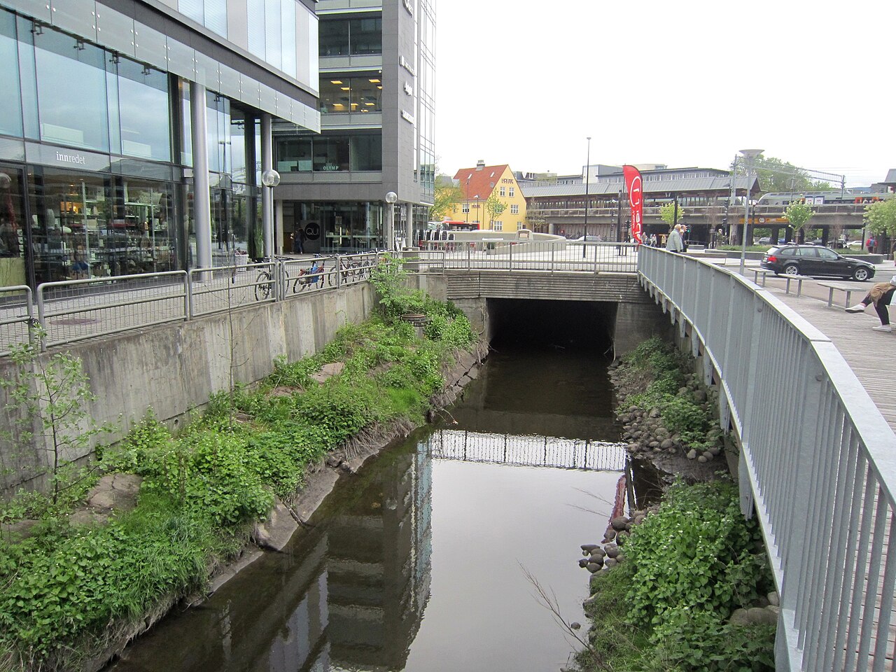 Narrow stream between vegetated banks, a modern building and a railing, running beneath a road bridge.
