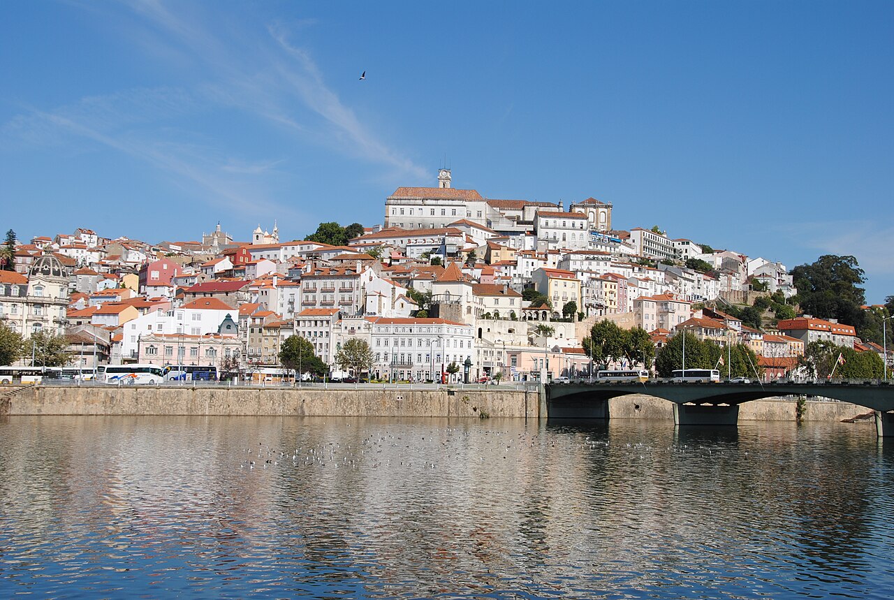 Wide river foreground, a bridge and buildings climbing the Coimbra hillside beneath a blue sky.