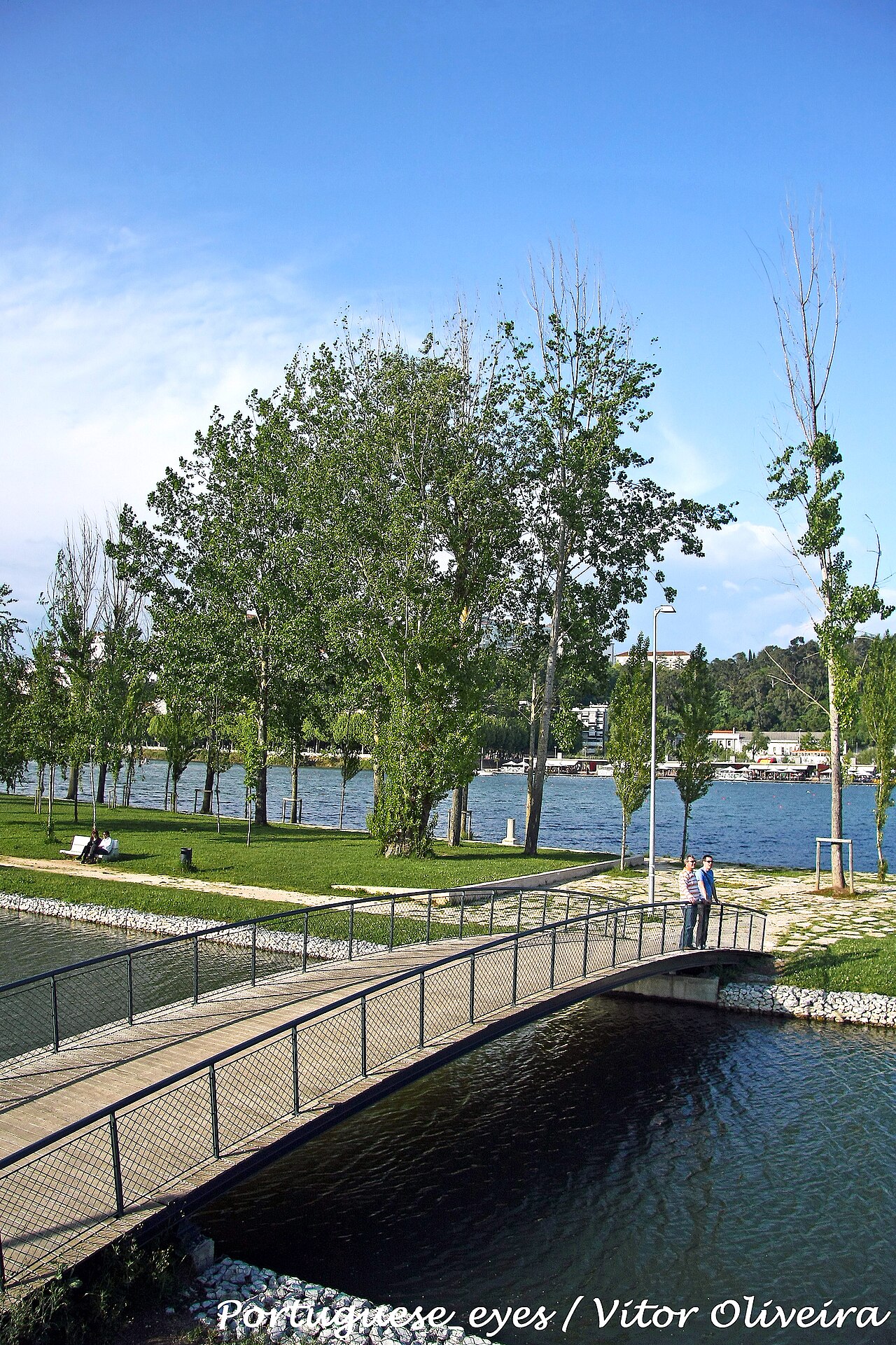 A pedestrian footbridge, trees and people beside the Mondego; the source photographer's watermark is retained.